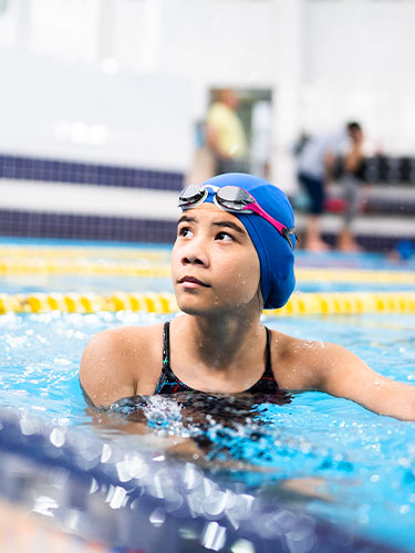 Girl with a blue swim cap on and goggles on her head, listens to her coach while in a pool.