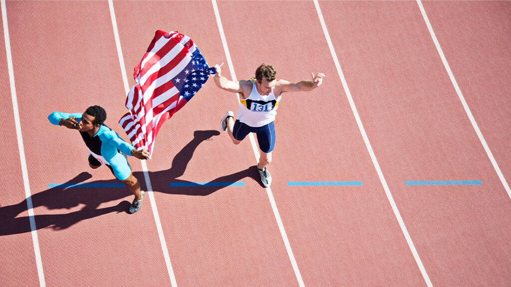 Male presenting adult track and field athlete running while holding the U.S. flag, with another adult athlete, wearing blue and black.