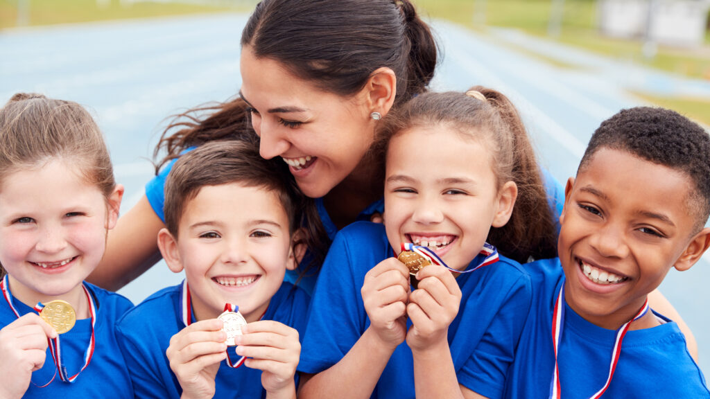 Four children wearing blue shirts show their small gold medals with red, white, and blue ribbons, being hugged by a smiling dark-haired woman.