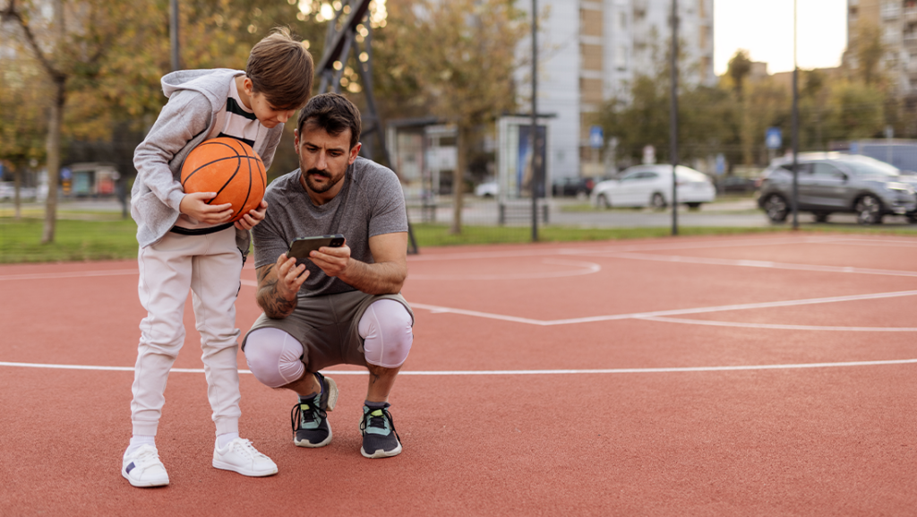 A father and his son stand on an outdoor basketball court, both focused on a mobile phone.