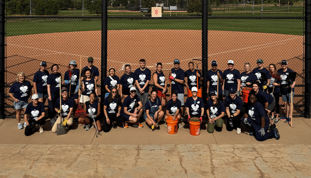 Group of around 30 employees volunteering for SafeSport Service Day 2025. Group is posing in front of a baseball field.