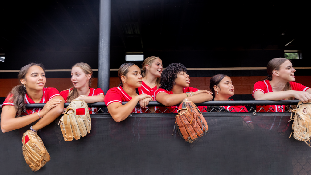 Teenage girls cheer on their softball teammates from dugout while holding softball gloves