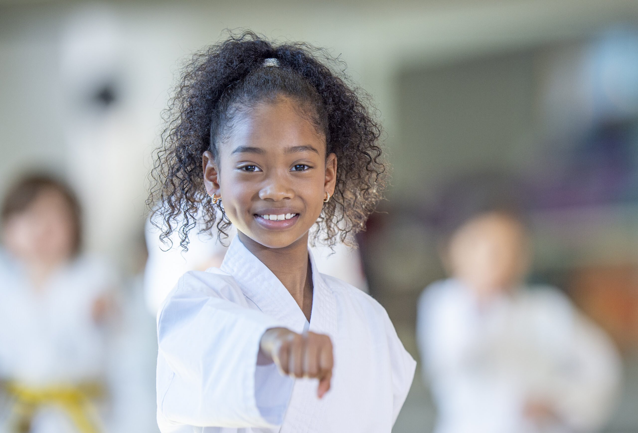 A young African American girl practices karate in a class setting and smiles at the camera.